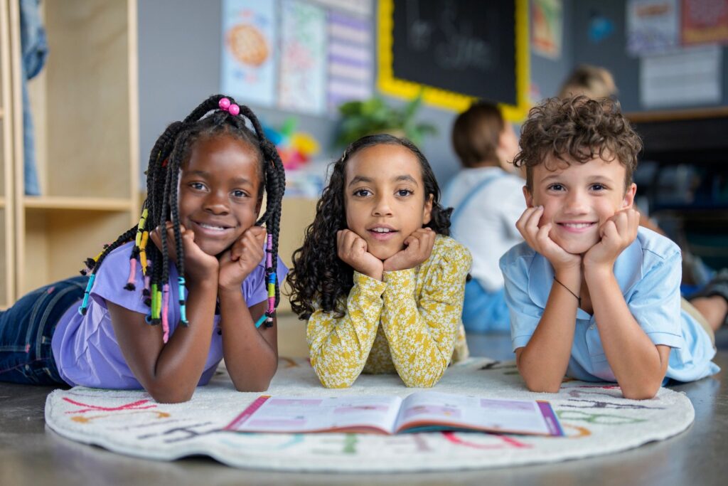 Three diverse children smiling and lying on the floor in a classroom.